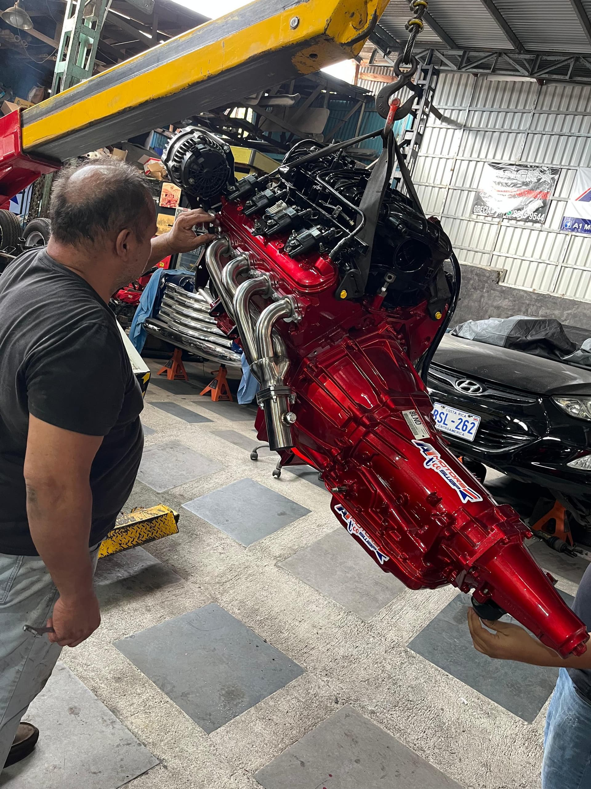 El LS2 6.0 y la caja 4L60 pintados en rojo colgando de la grúa, con múltiples tipo header en acero inoxidable y calcomanías de A1 Motors Tico Swap