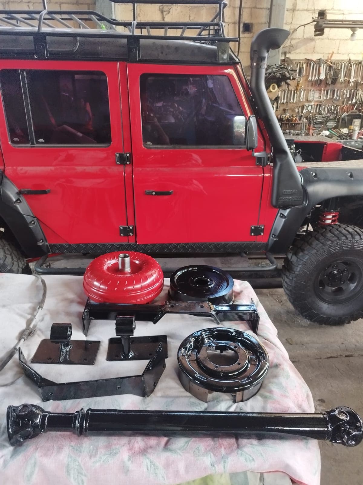 Convertidor de torque, cardán, soportes de motor y platos de freno pintados sobre la mesa, con el Land Rover Defender rojo al fondo