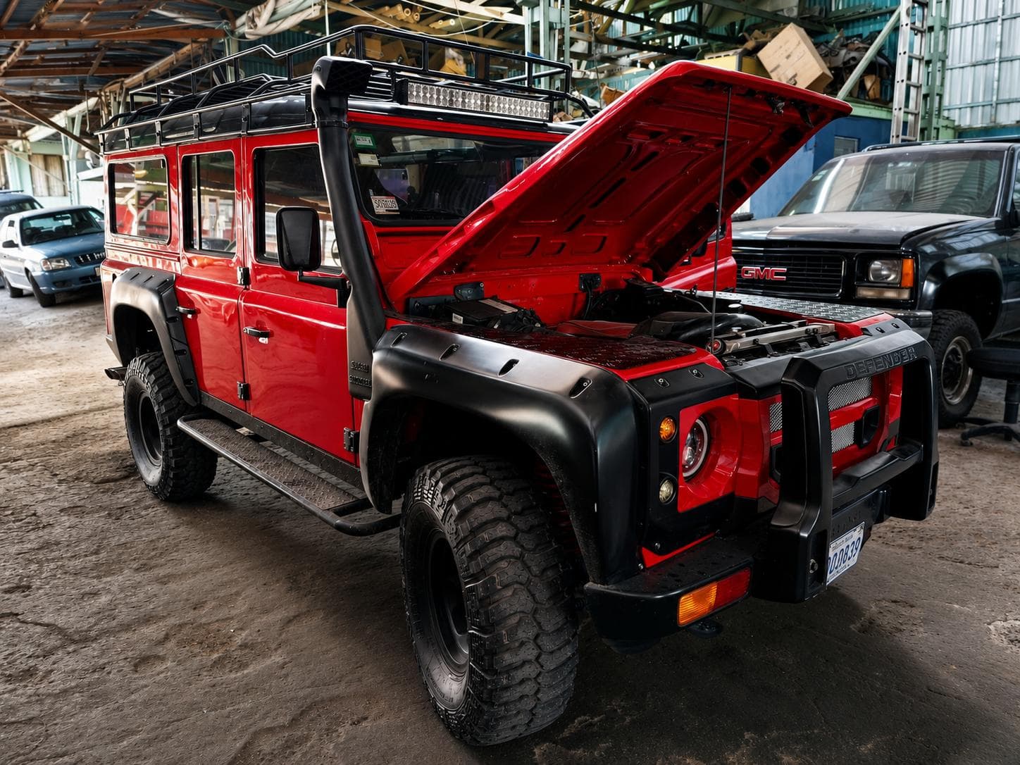 Land Rover Defender rojo con parrilla de techo, snorkel y llantas de barro, con el capó abierto en el taller de A1 Motors Tico Swap